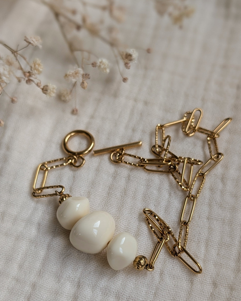 Bracelet PAULE sur fond gaze de coton déplié. Chaîne trombone en acier inoxydable doré et perles résine couleur ivoire. Création artisanale La Tête dans les Nétoiles