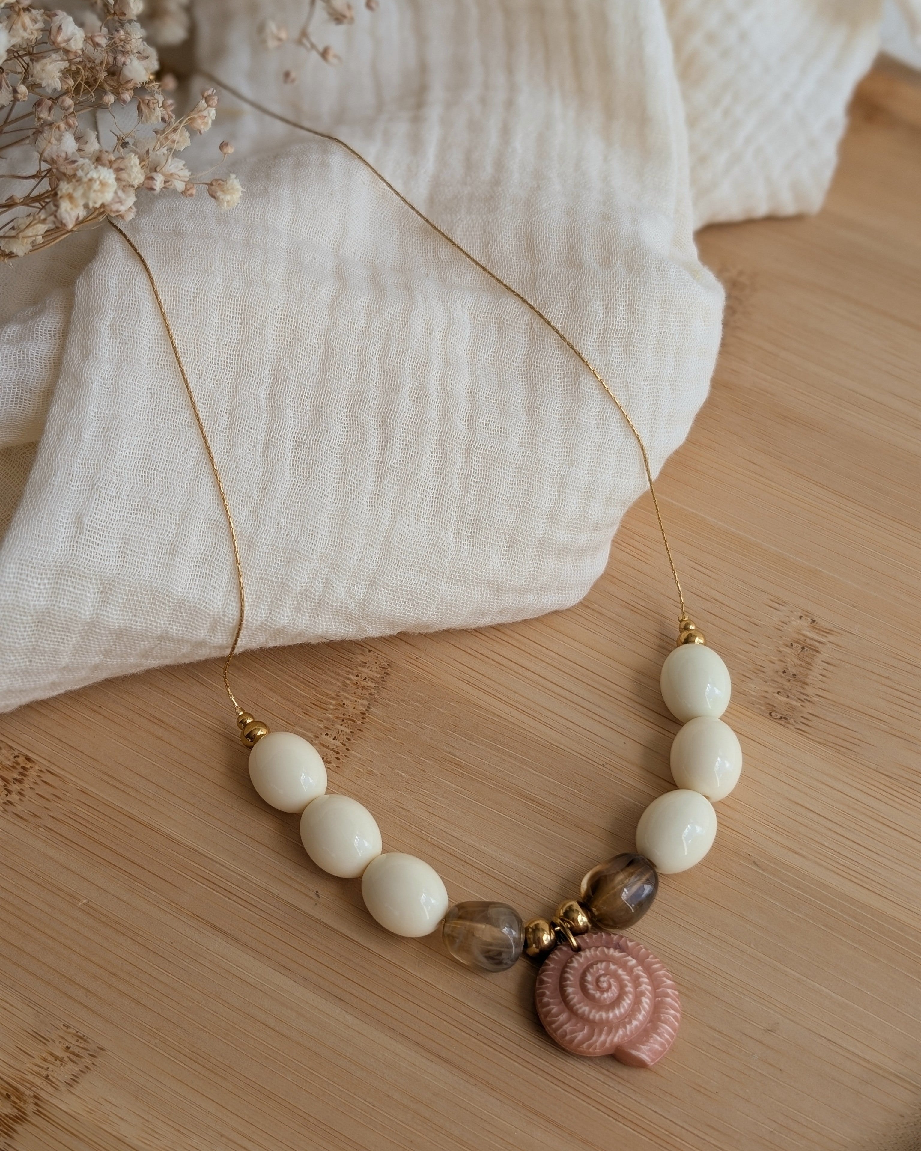 Collier Azura en entier sur plateau en bois. Perles crème et marron forme olive et pendentif coquillage ammonite marron clair. Perles en résine et maille en acier inoxydable. Création artisanale La Tête dans les Nétoiles 