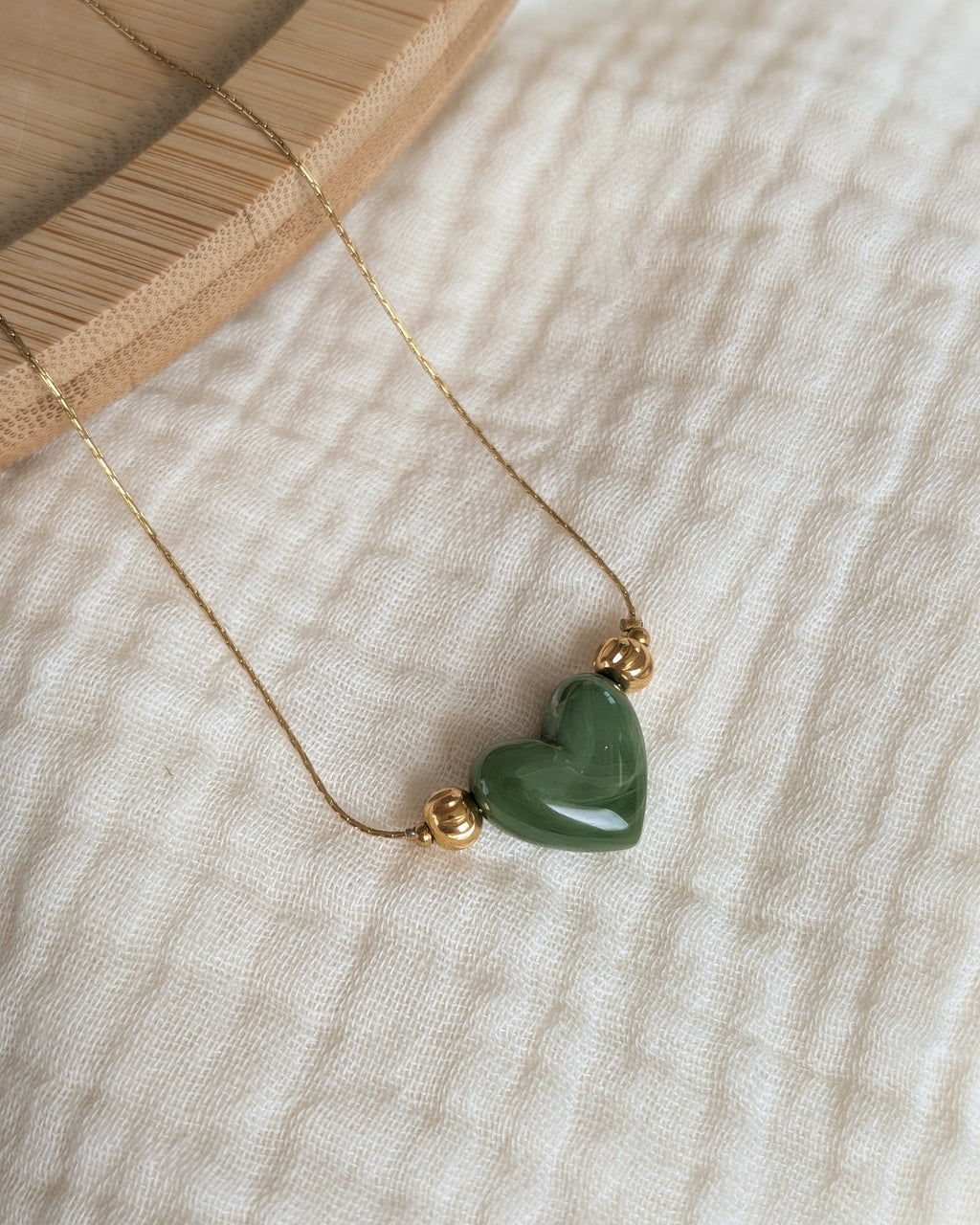 Collier Aimée perle coeur en résine vert kaki et acier inoxydable doré. Posé sur plateau en bois et gaze de coton écru. Créations faits main La Tête dans les Nétoiles
