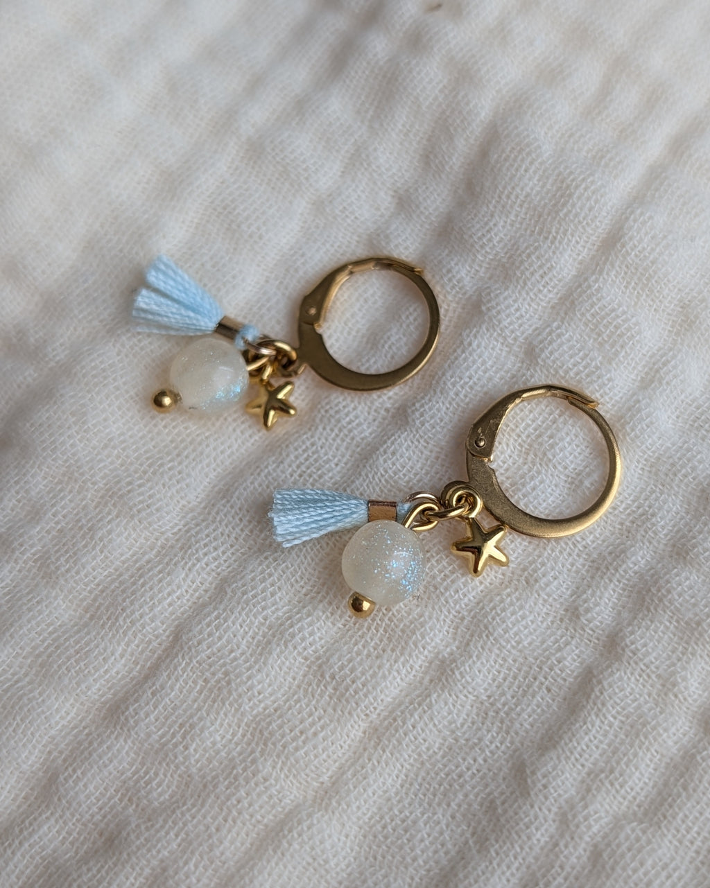 Gros plan boucles d'oreilles enfant créoles avec étoile, perle irisée et pompon bleu. Créole en acier inoxydable doré. Création artisanale La Tête dans les Nétoiles