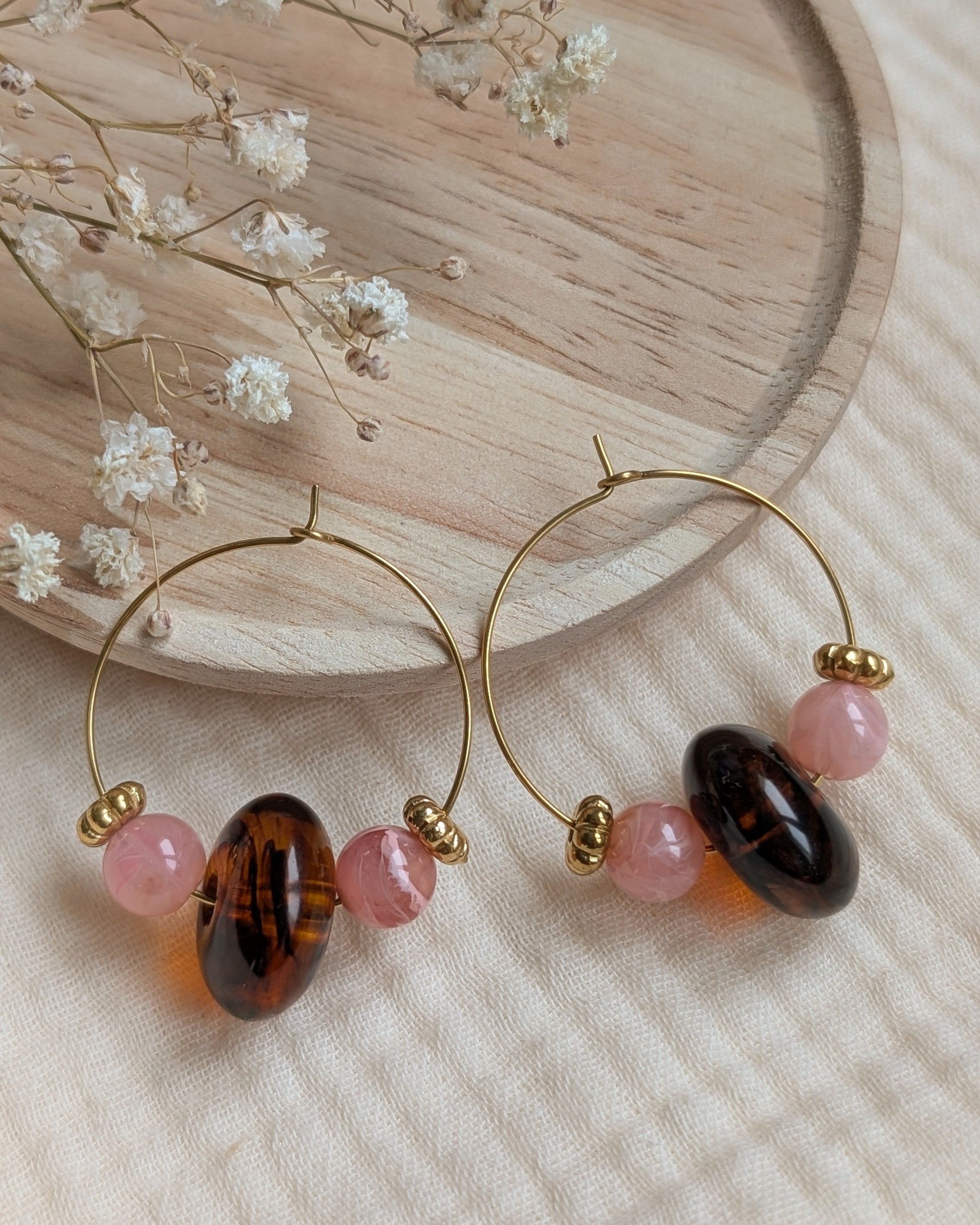 Boucles d'oreilles créoles Elise en acier doré avec perles rose poudré et perle centrale ambre foncé sur tissu beige. Création artisanale La Tête dans les Nétoiles