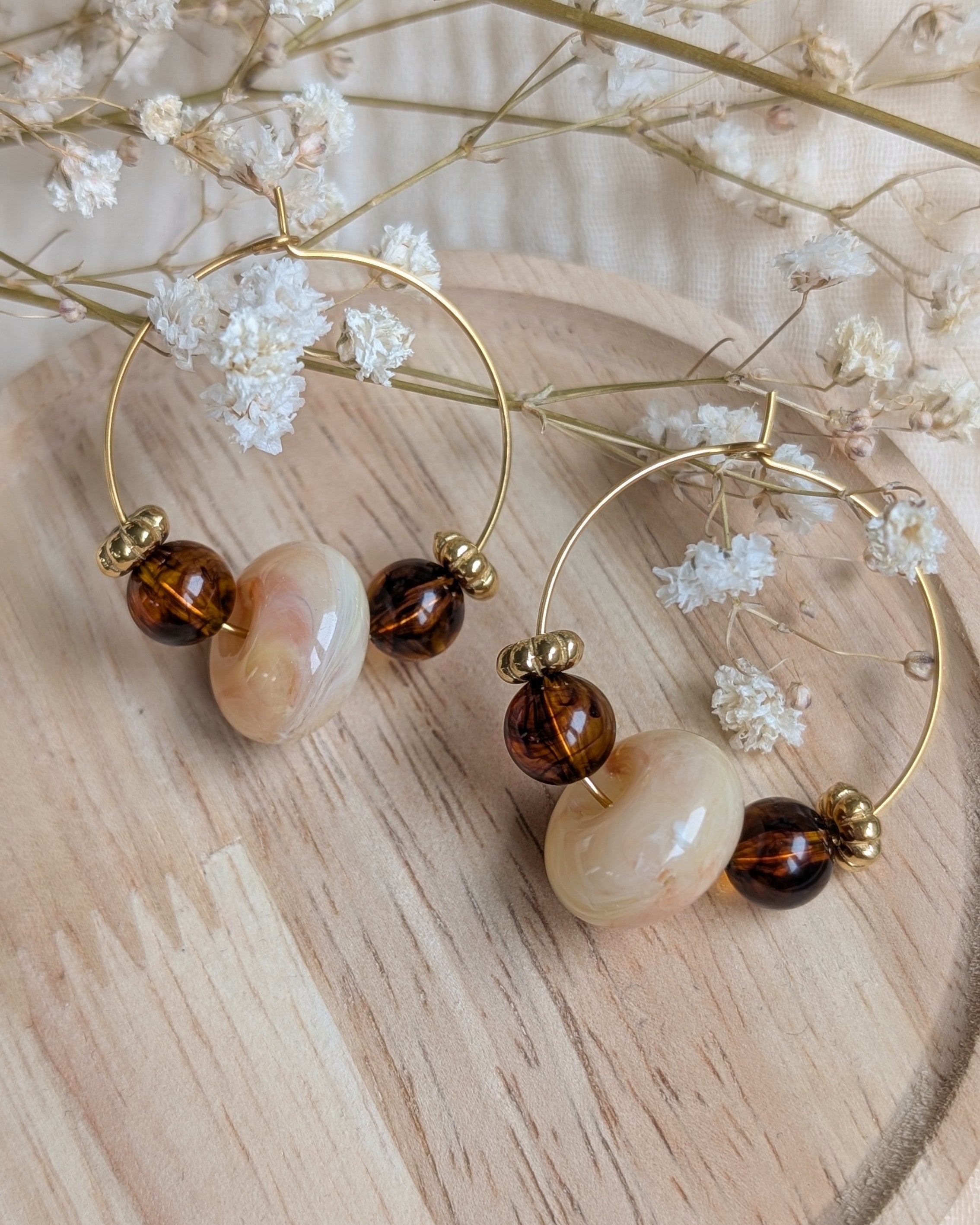 Paire de boucles d'oreilles créoles dorées artisanales Elise avec perle centrale marbrée crème et perles ambrées sur plateau en bois. Création La Tête dans les Nétoiles