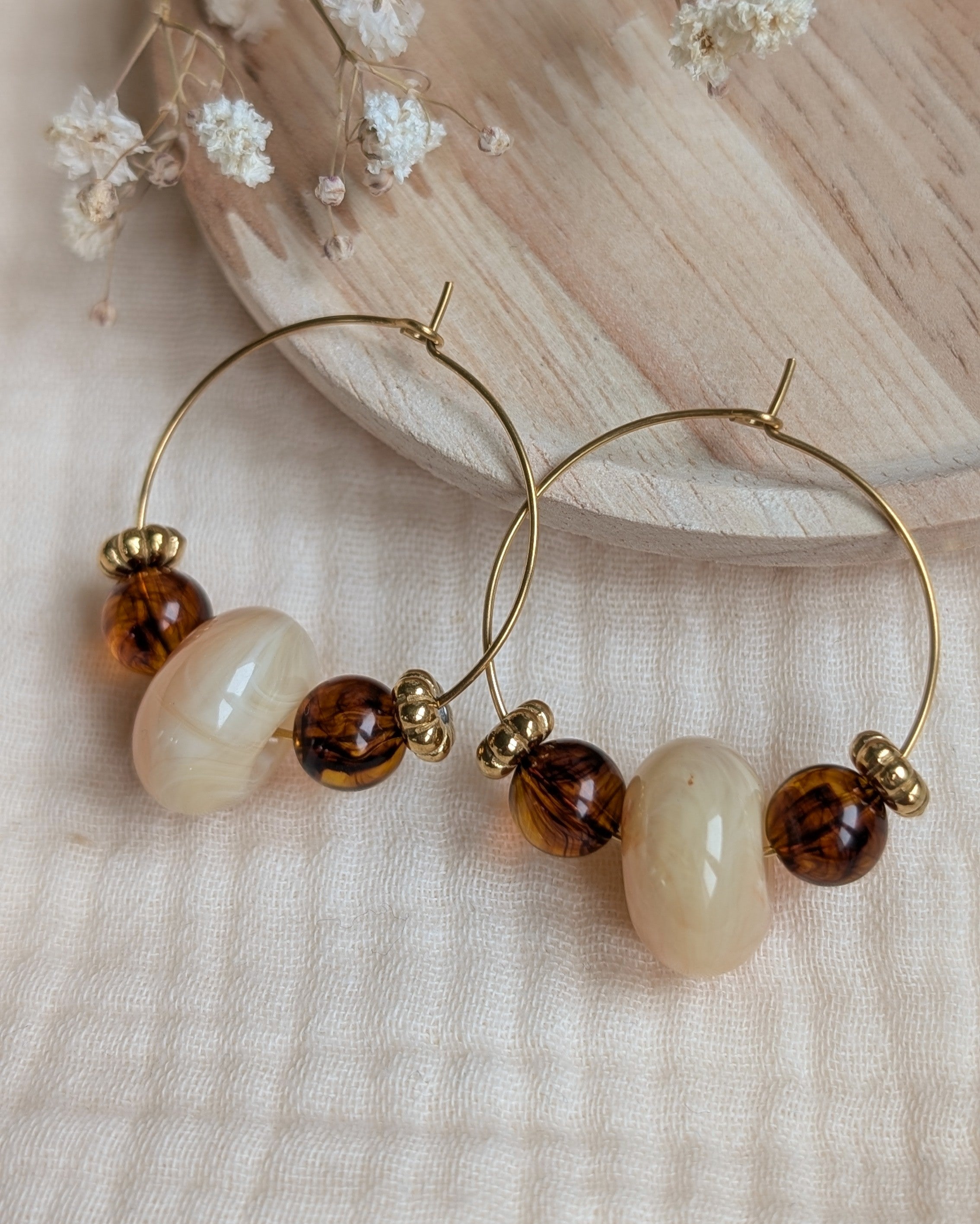 Paire de boucles d'oreilles créoles dorées en acier inoxydable créations artisanales Elise avec perle centrale marbrée crème et perles ambrées sur plateau en bois. La Tête dans les Nétoiles