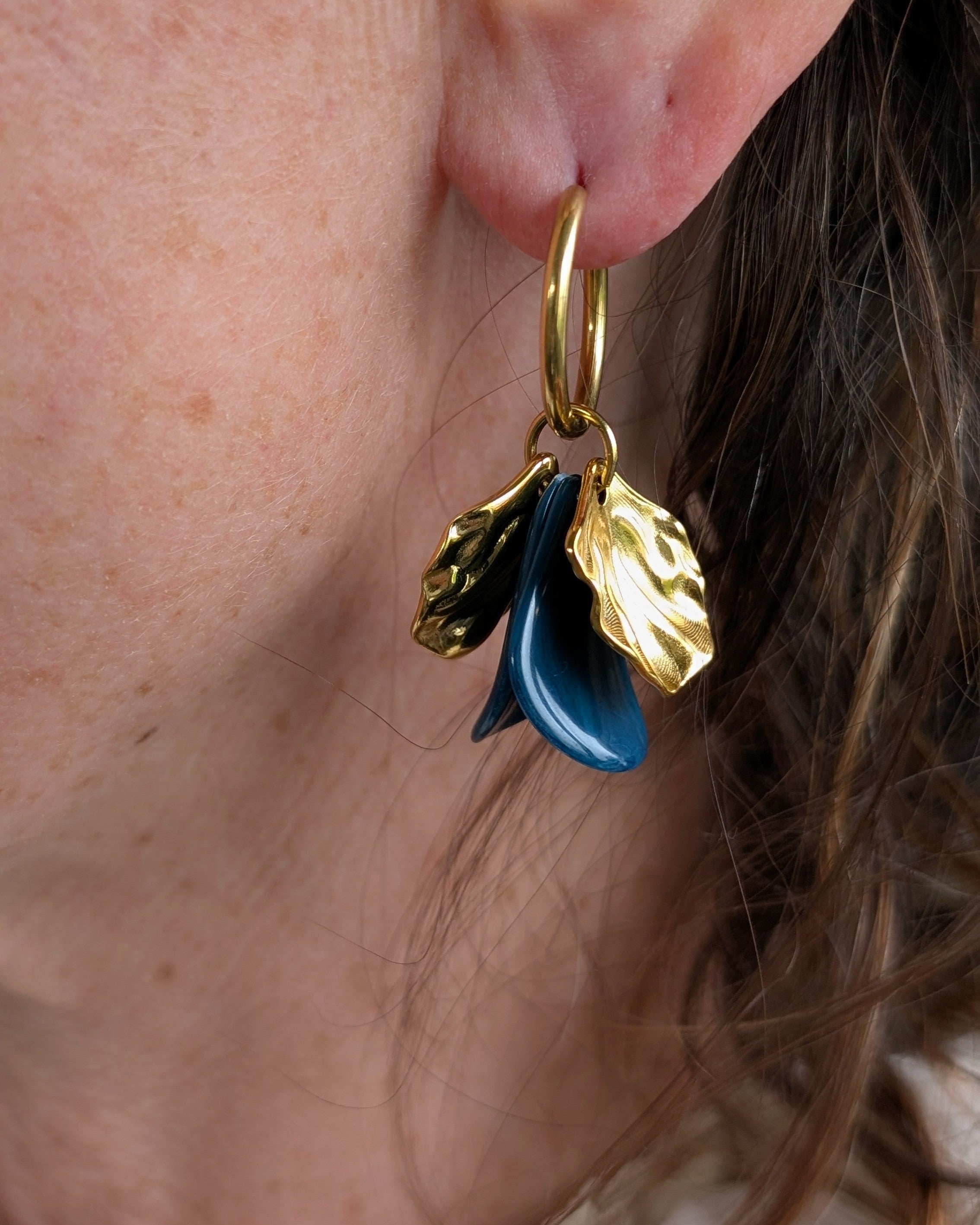 Boucles d'oreilles créoles Sora avec pétales bleus paon en résine et feuilles dorées sculptées, portées par une femme. Créoles en acier inoxydable.  Création artisanale La Tête dans les Nétoiles