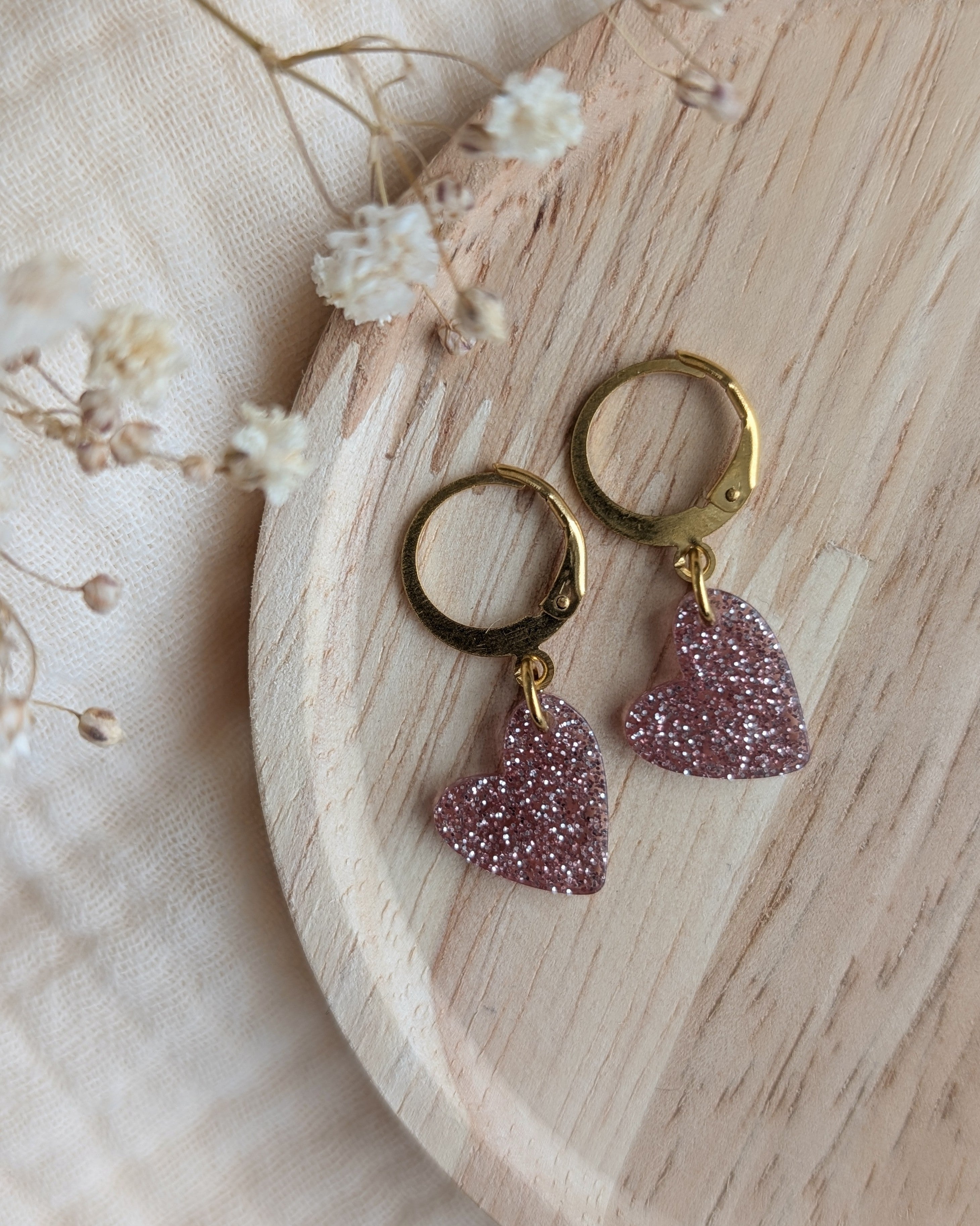 Détail des boucles d'oreilles petites filles cœur rose pailleté posée sur un support en bois. Création artisanale La Tête dans les Nétoiles