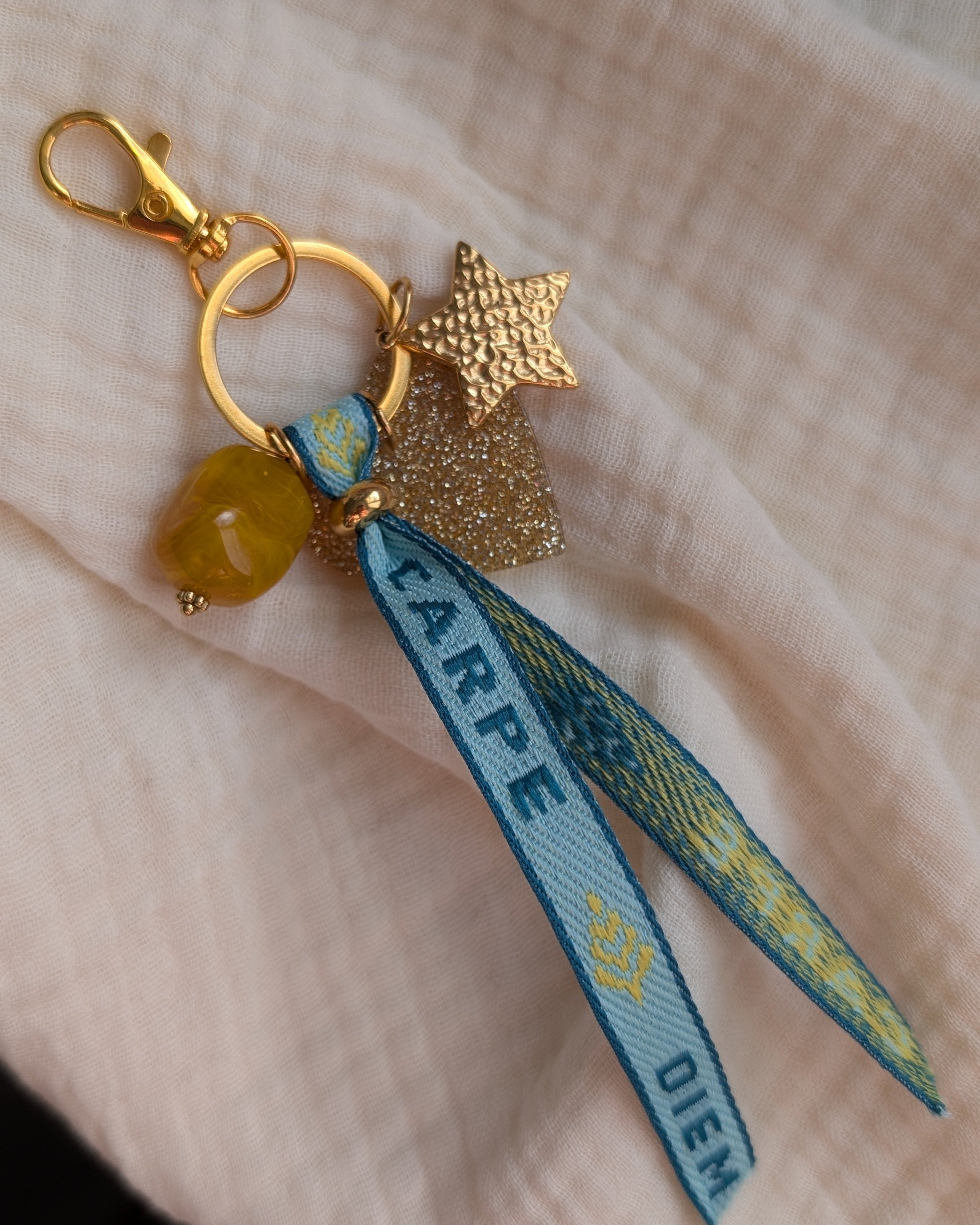 Porte clés ou bijou de sac ruban tissé carpe diem. Pendentif perle en résine jaune, pendentif coeur doré à paillettes et étoile en acier inoxydable. 
Création La Tête dans les Nétoiles