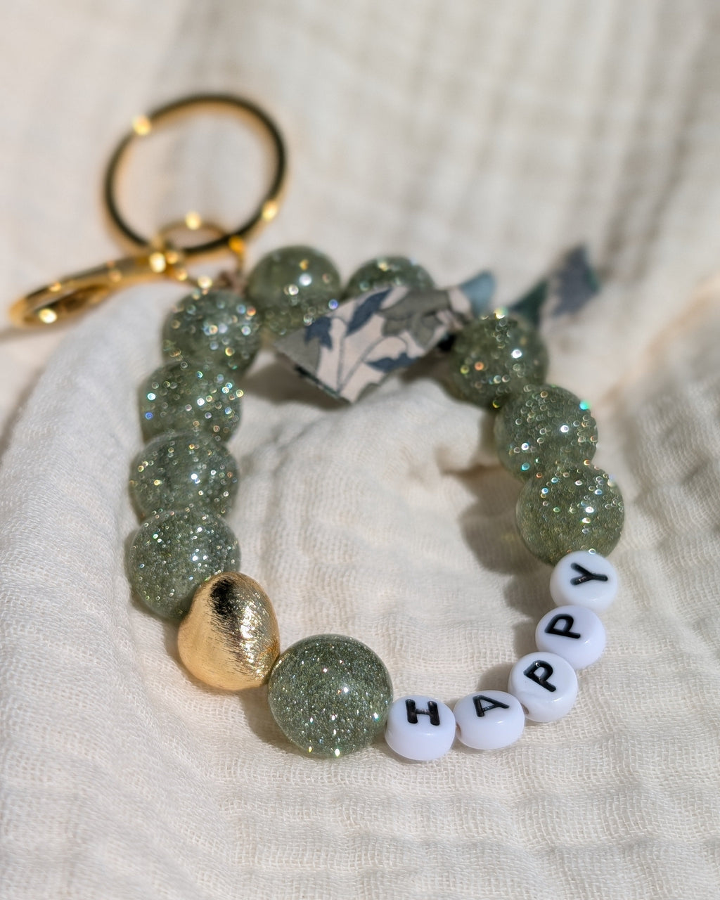 Porte clés et bijoux de sac liberty glowy vert clair paillettes posé sur gaze de coton. Mot happy à personnaliser. Créations artisanales La Tête dans les Nétoiles