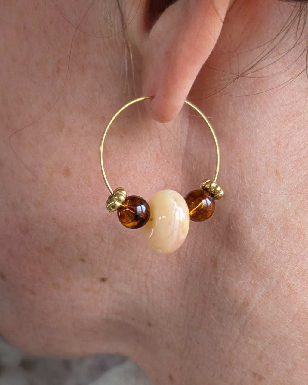 Gros plan d'une boucle d'oreille créole Elise portée, révélant la transparence des perles ambrées et beige marbrées. Création artisanale La Tête dans les Nétoiles