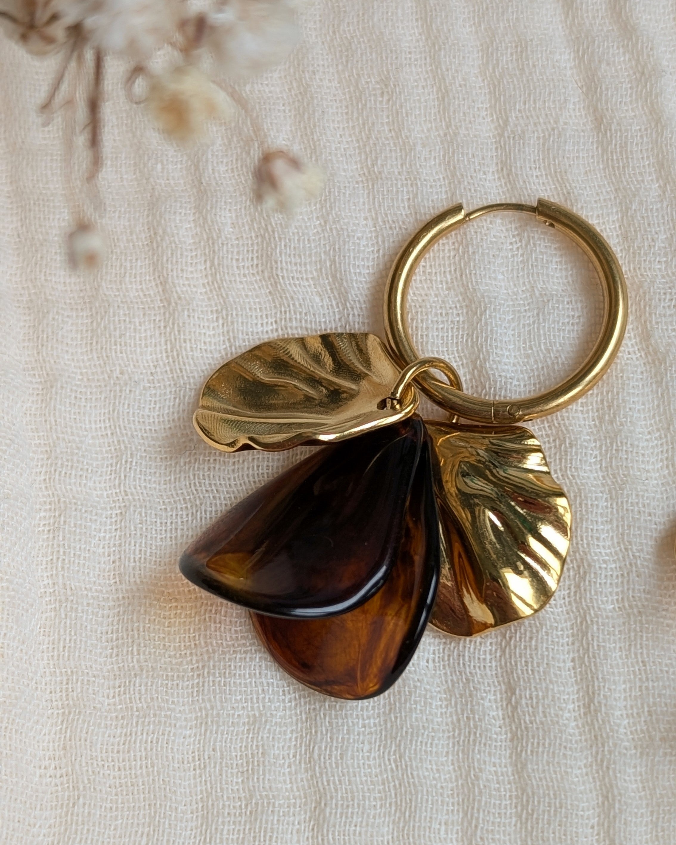 Détail d'une boucle d'oreille Sora ambrée posée sur un tissu de gaze de coton blanc, pétales en acétate marbré et feuilles dorées texturées. Création artisanale La Tête dans les Nétoiles