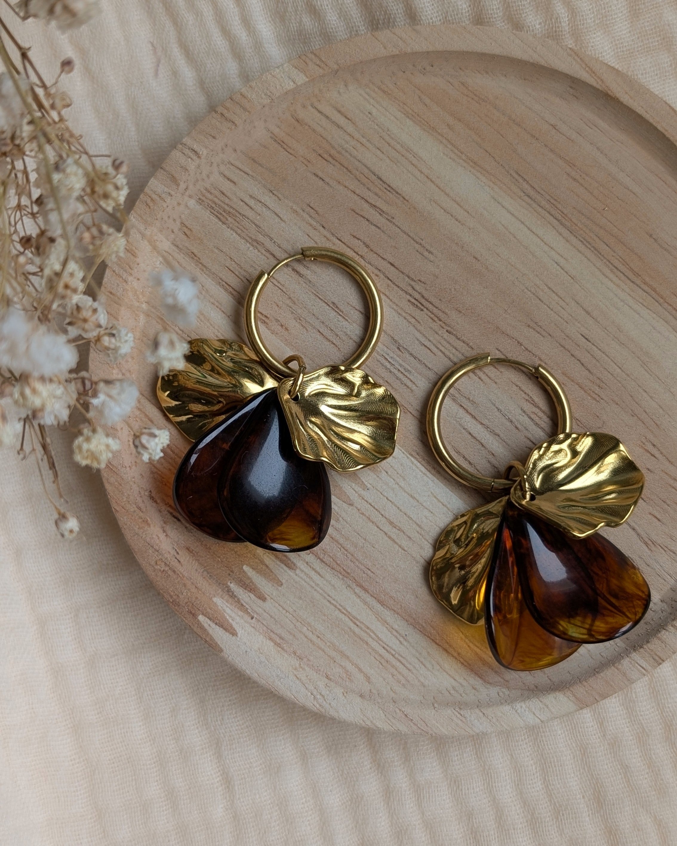 Boucles d'oreilles Sora ambrées et dorées posées sur un plateau en bois, détails des pétales en acétate miel et feuilles sculptées. Création artisanale La Tête dans les Nétoiles