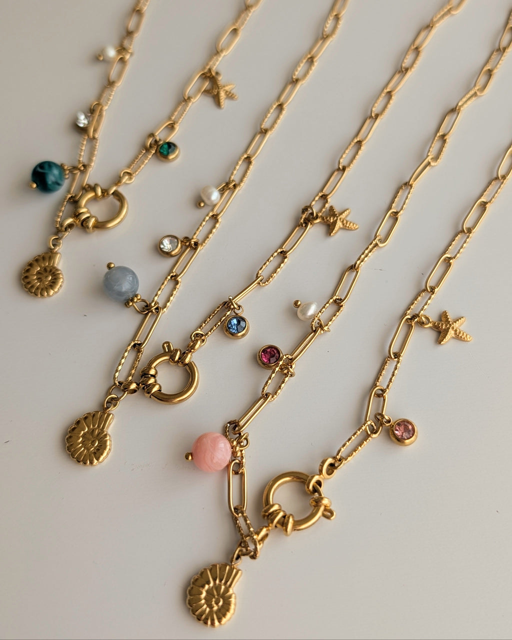 Trio de colliers Océane chaine maille trombone posés sur table en bois blanc. Détails sur les 3 coloris. Charms marins. pendentifs ammonite et étoile de mer. Créations artisanales La Tête dans les Nétoiles
