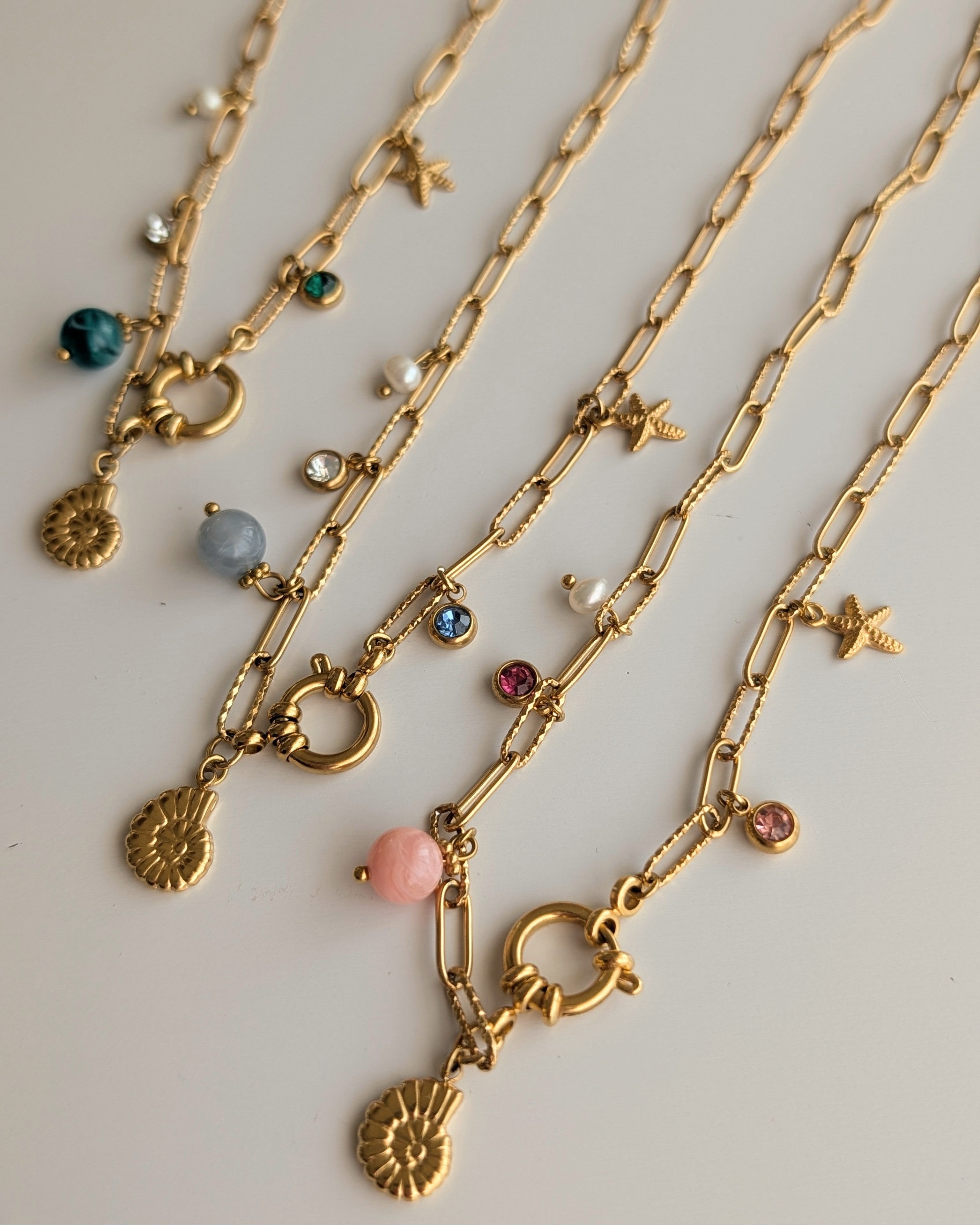 Trio de colliers Océane chaine maille trombone posés sur table en bois blanc. Détails sur les 3 coloris. Charms marins. pendentifs ammonite et étoile de mer. Créations artisanales La Tête dans les Nétoiles