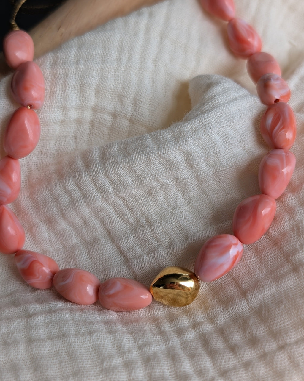 zoom sur la perle dorée du collier Louison colori rose corail. Posé sur gaze de coton. Création La Tête dans les Nétoiles